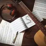Music sheet, headset, a guitar and a pen on a table, all of which are materials used when studying at the best colleges for music.