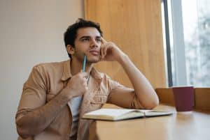 A male student seated by the window is holding a pen and is lost in his thoughts.