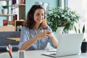 A woman is smiling while wearing headphones and watching on her laptop.