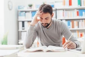 A man in the library reading a book and writing down notes at the same time.