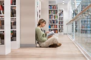 A female student sitting in lotus position on the library floor near bookshelves.