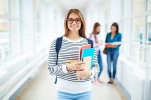 A female student wearing glasses and carrying books is standing in the hallway.
