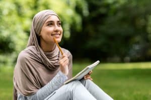 A girl in hijab writing essay while sitting on the grass.