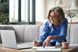 A woman sitting on a couch and writing with coffee and laptop beside her.