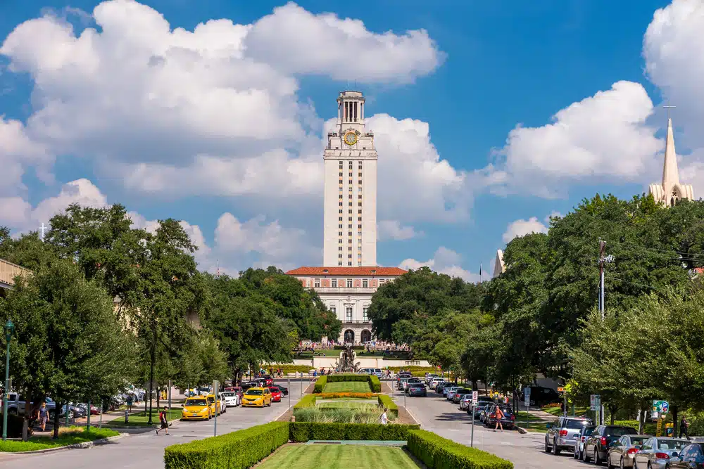 UT Austin's tall white building with trees and vehicles on both sides.