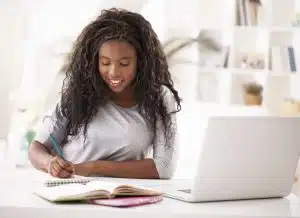 a student writing in her notes on a table with a white laptop