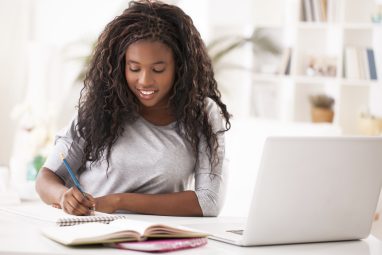 a student writing in her notes on a table with a white laptop