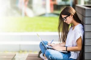 Female student typing an essay while sitting on the pavement.