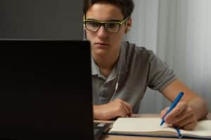 Male student writing in his notebook while looking at the laptop.