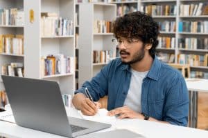 Male student using a laptop for college application in a libaray.