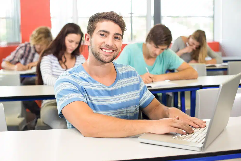 Male student typing in a laptop in a classroom.