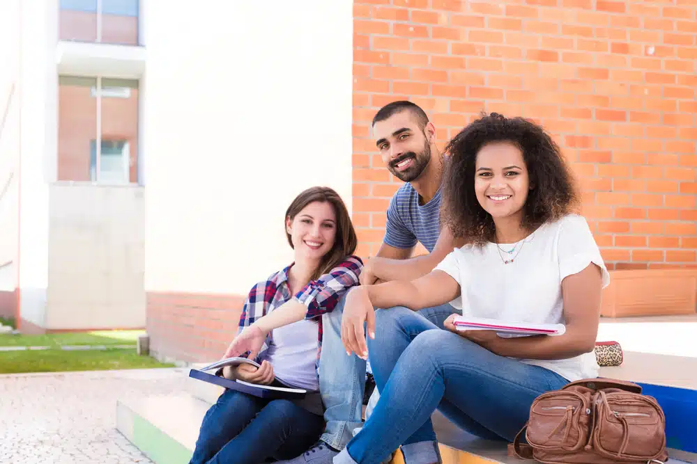 Three students sitting near a building and smiling at the camera.