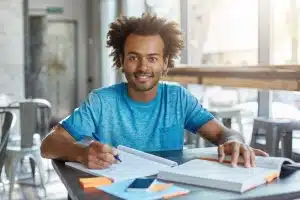 Male student sitting on a table with books while smiling at the camera.