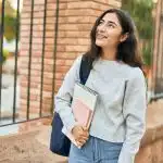A young woman carrying notebooks walks on campus, representing the choice between UT Austin vs University of Michigan.