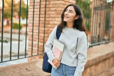 A young woman carrying notebooks walks on campus, representing the choice between UT Austin vs University of Michigan.
