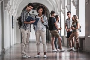 A male and female student standing in the middle of the hallway, talking to each other while the female student is looking through the notebook that the male student is holding