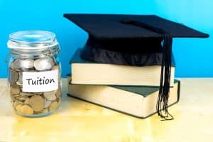 A jar of coins beside a book and a graduation cap.