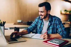 a male phd student smiling as he writes something on his notebook