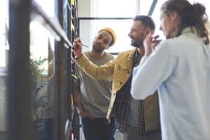 3 male standing adjacent to each other while the person in the middle is writing on the glass board while the rest of them pays attention