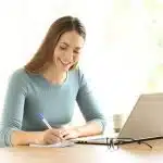 A woman smiling in front of a laptop while taking notes as she compares Northeastern vs UC Irvine.