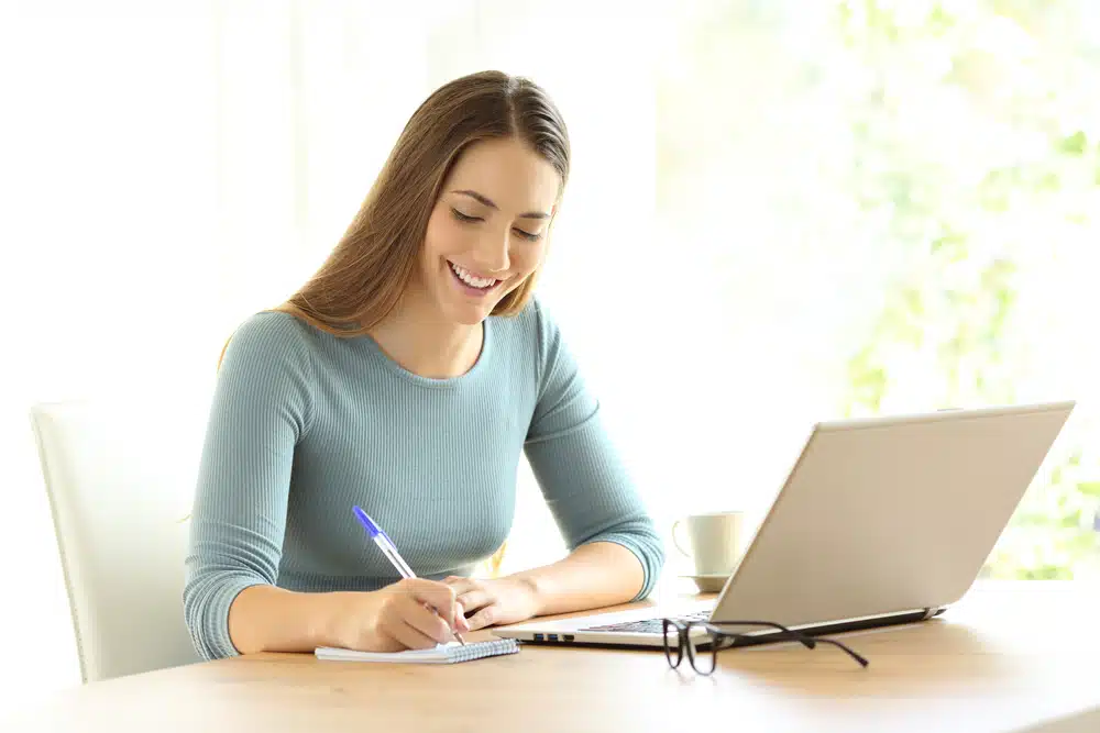 A woman smiling in front of a laptop while taking notes as she compares Northeastern vs UC Irvine.