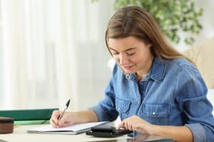 A female student writing on a table while using a calculator.