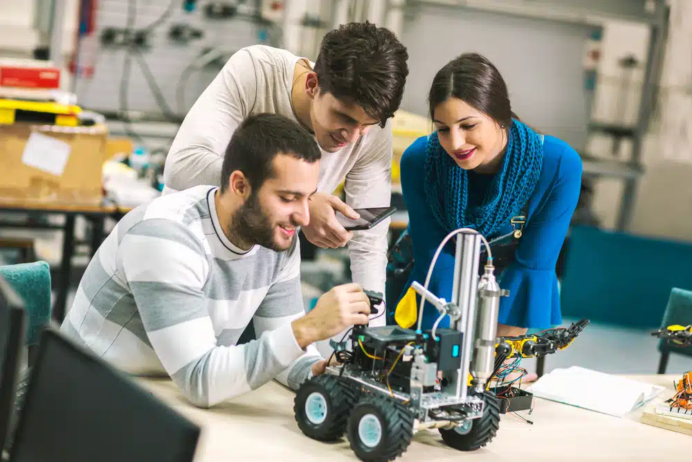 a female and two male mechanical engineering students gathered around and studying a tiny robotic vehicle