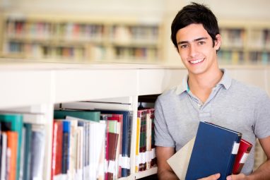 A smiling student holds books in a library while considering University of Michigan vs Boston College.
