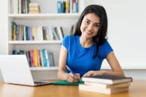 a student writing on her notebook and looking at the camera