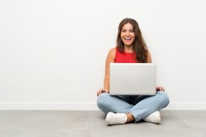 A students using her laptop while sitting next to a wall.