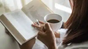 Asian woman holding cup of coffee and reading a book beside the window after get up in morning.