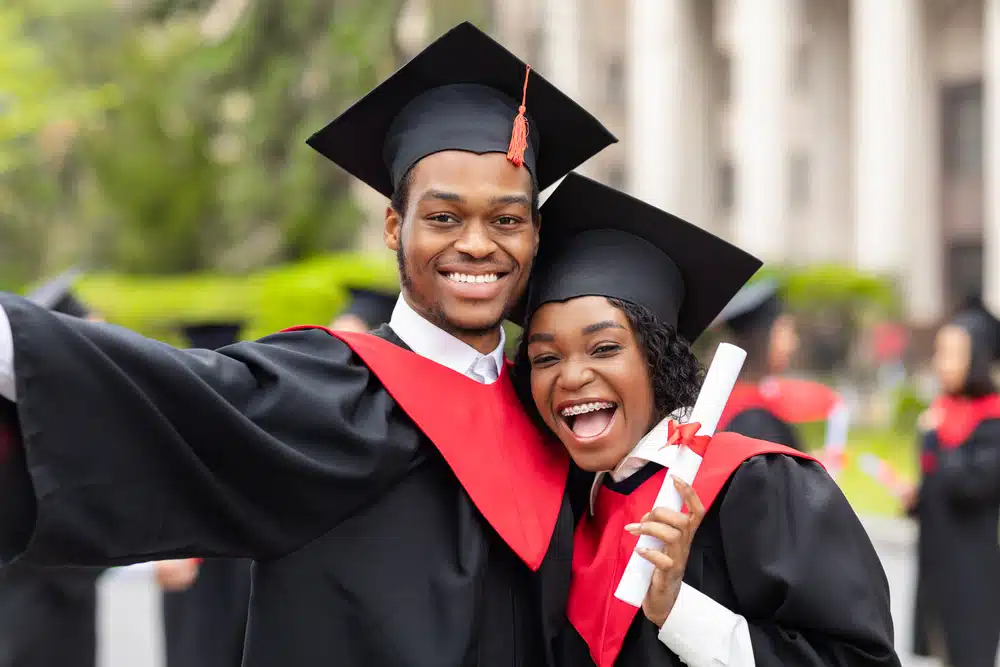 a male and female student wearing their graduation gown, looking happy while looking at the camera