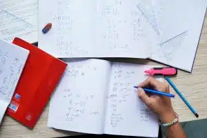 an image of a hand writing unto a notebook while other sheets of paper are sprawled on the table