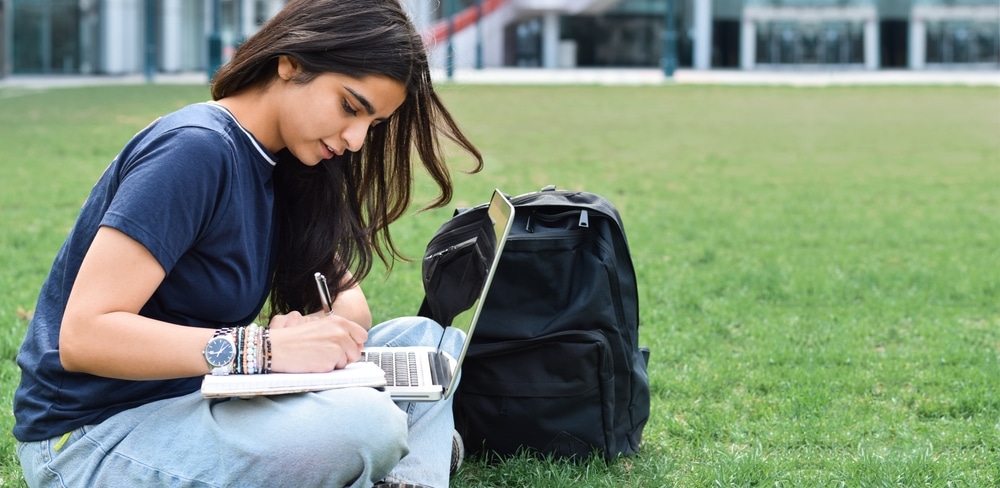 Young girl student sitting on grass outside using laptop computer