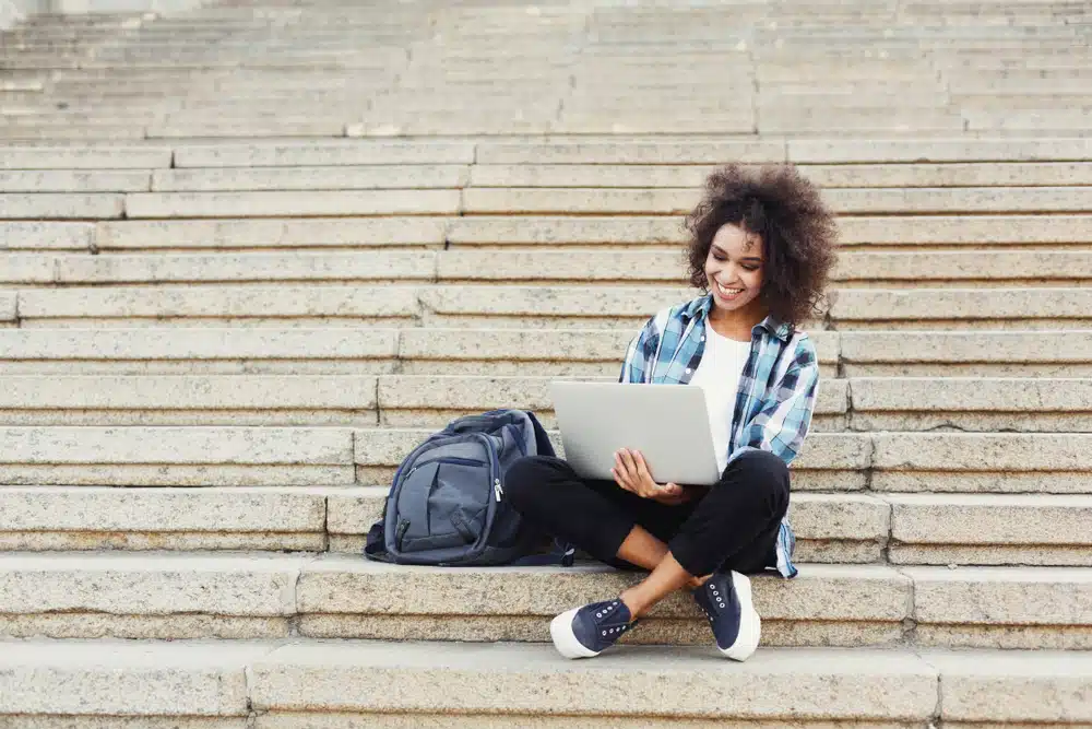 Smiling african-american student girl sitting on stairs working on laptop