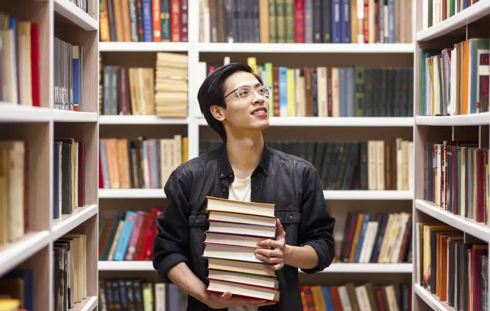 A bookworm smiling while holding a pile of books in the library.