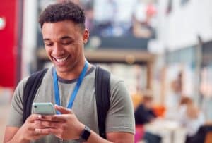 Smiling Male College Student Checking Mobile Phone In Busy Communal Campus Building