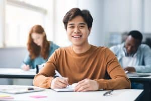 Portrait of a smiling Asian male student sitting at a desk in a classroom at the university