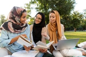 Photo of happy young Arabian women students using laptop computer and holding books in park