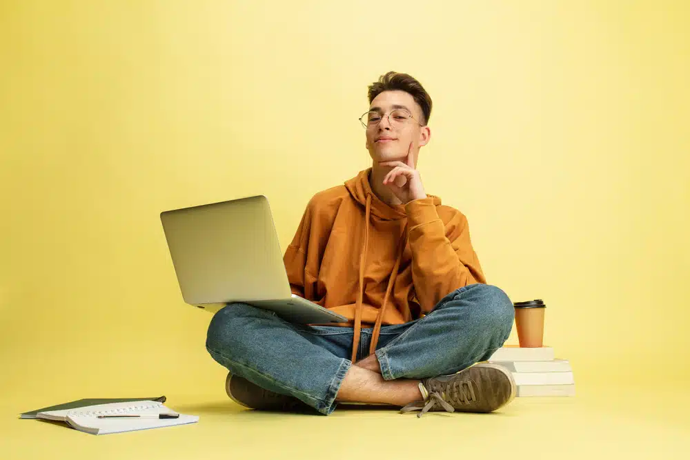 One young smiling caucasian man, student in glasses sits on floor with laptop isolated on yellow studio background.