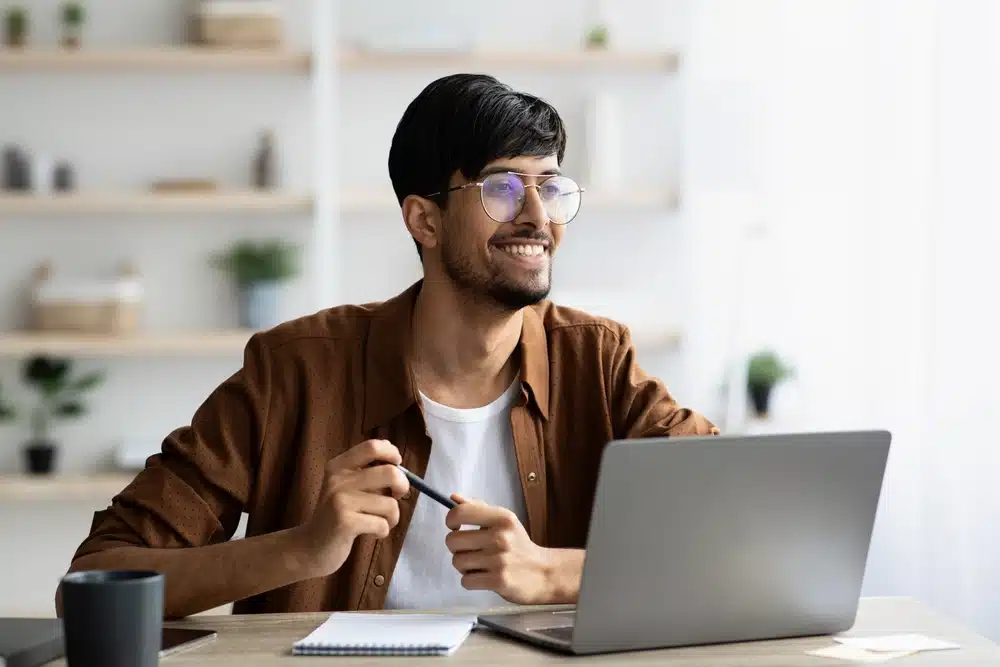 Excited middle eastern young man in casual sitting at workdesk, working on laptop at office