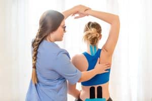 Young female patient wearing kinesio tape on her back and neck exercising with a professional physical therapist.