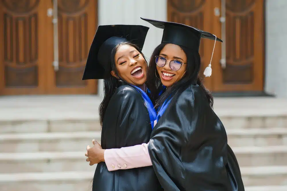 two young ladies in graduation costumes posing at camera at university campus, holding diplomas, laughing and hugging