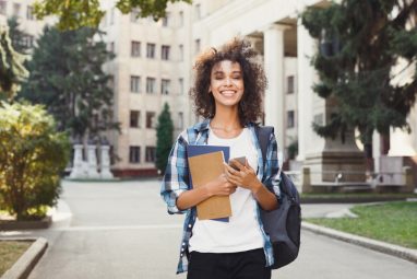 A smiling student holds books and a phone on campus, possibly comparing USC vs NYU.