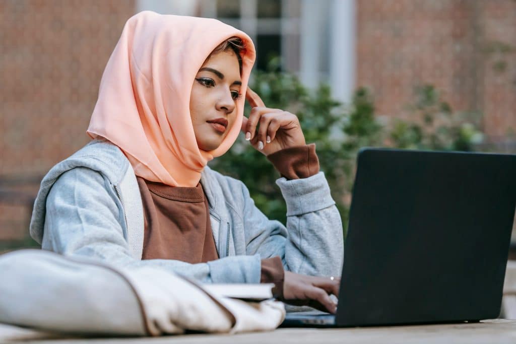 A young woman wearing a peach hijab studies on her laptop outdoors while considering UPenn vs Berkeley.