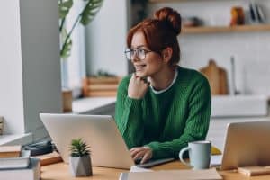 lady in green sweater looking outside with laptop and mug