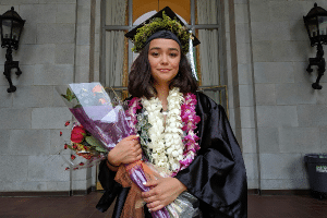 A graduate posing for the camera holding flowers while wearing lei flower necklaces