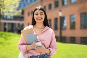 Portrait of happy student girl with cellphone and workbooks walking in college campus and smiling at camera