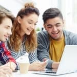 Three college students smiling as they work together on a laptop while discussing Dartmouth GPA requirements.