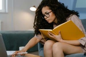A woman holding a yellow notebook crouched while reaching for her laptop to scroll